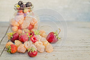 Multicolored raspberries in a glass jar with strawberries on background