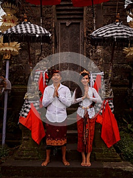 Multicultural couple standing near the temple in Bali. Hands in namaste. Caucasian wife and Balinese husband wearing traditional
