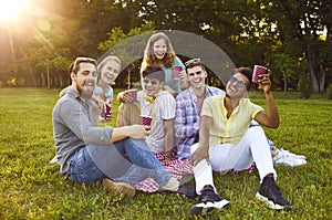 Multicultural friends having fun drinking from paper cups sitting on green lawn in park.