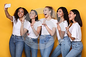 Multicultural Ladies Making Selfie On Phone Gesturing V-Sign, Yellow Background