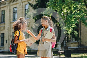 Multicultural schoolgirls holding books and speaking while standing in schoolyard