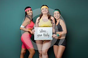 Multicultural sportswomen smiling, looking at camera and showing placard with love your body lettering