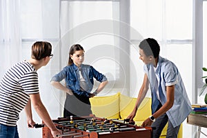 multicultural teen boys playing table football and female friend standing with hands akimbo