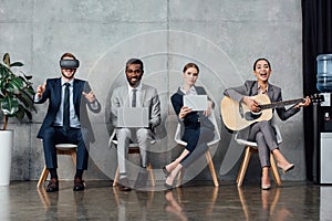 Multiethnic businesspeople sitting on chairs, using digital devices and playing acoustic guitar in photo