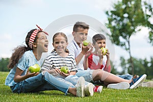 Multiethnic children eating green apples while sitting together on green grass