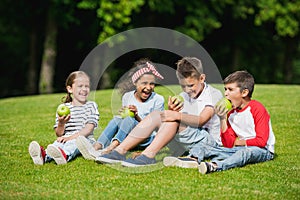 Multiethnic children eating green apples while sitting together on green grass