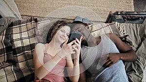 Multiethnic couple lying on bed and using smartphone. Man and woman happy together. Male and female laughing, smiling.