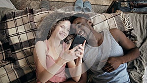 Multiethnic couple lying on bed and using smartphone. Man and woman happy together. Male and female laughing, smiling.