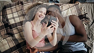 Multiethnic couple lying on bed and using smartphone. Man and woman happy together. Male and female laughing, smiling.