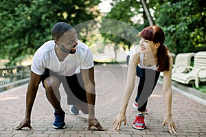 Multiethnic couple in sport clothes on starting line preparing to run in the park, smiling and looking each other