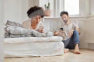 Multiethnic couple watch video via social networks, hold modern cellulars, wife lies in bed, husband sits near at floor