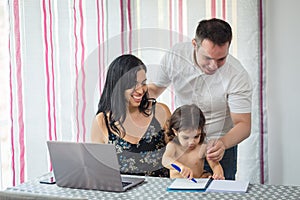 Family gathered at the dining room table, mother at the laptop, father standing hugging them and baby girl painting in a notebook