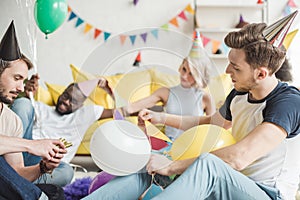 multiethnic friends in party hats sitting on floor with balloons in decorated