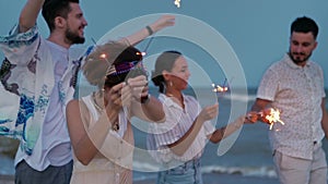 Multiethnic friends walking, dancing, having fun on night party at seaside with sparklers lights in hands on Fourth of