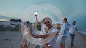 Multiethnic friends walking, dancing, having fun on night party at seaside with sparklers lights in hands on Fourth of