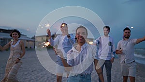 Multiethnic friends walking, dancing, having fun on night party at seaside with sparklers lights in hands on Fourth of