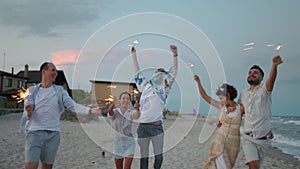 Multiethnic friends walking, dancing, having fun on night party at seaside with sparklers lights in hands on Fourth of