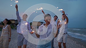 Multiethnic friends walking, dancing, having fun on night party at seaside with sparklers lights in hands on Fourth of