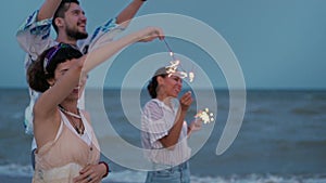 Multiethnic friends walking, dancing, having fun on night party at seaside with sparklers lights in hands on Fourth of