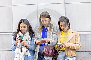 Multiethnic Group of beautiful young female friends standing against a wall using mobile smartphones