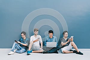 Multiethnic group of four men using smartphone, laptop computer, digital tablet together with copy space on blue wall
