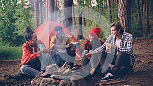 multiethnic group of friends girls and guys are sitting in forest around fire with drinks clinking glasses and smiling