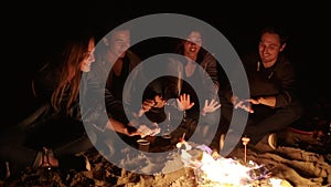 Multiethnic group of people sitting by the bonfire at the beach and warming up their hands in the evening, talking and