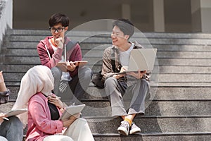 A multiethnic group of university students is studying together while sitting on steps near the campus and communicating