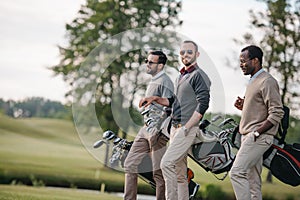 Multiethnic men holding bags with golf clubs and walking on golf course