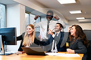 Multiethnic office team rejoices around computer, happy African exec raises fists in victory, coworkers cheer, man photo
