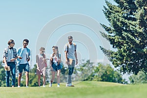 multiethnic teenage students with books and skateboard talking while walking together in park