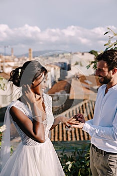 Multiethnic wedding couple - African-American bride and Caucasian groom. Groom puts a ring on brides finger. Destination