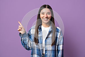 Multiethnic woman with attractive smile pointing with index finger up looking in camera against purple background