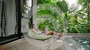 Multiethnic woman relaxes with laptop by poolside on weekend. Comfortable beanbag, lush tropical garden view. Casual