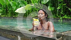 Multiethnic woman relaxes in pool, sips fresh cocktail among rich plants. Satisfied smile, leisure day, enjoys exotic