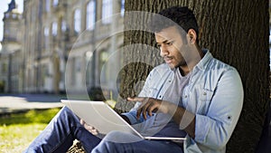 Multinational young man sitting under tree, looking at laptop reading job offer