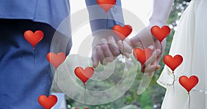 Multiple heart balloons floating against mid section of newly married couple holding pinky finger