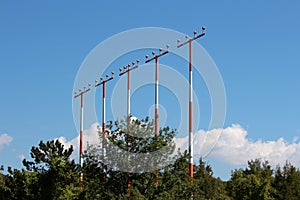 Multiple high metal red and white poles with airport runway guiding lights surrounded with dense trees