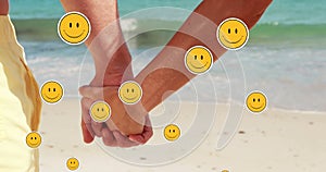 Multiple smiling face emoji floating against mid section of couple holding hands at beach