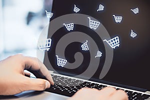 Multiple white shopping carts icons with hands touching the laptop keyboard in the background. Online shopping and purchase