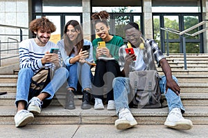 Multiracial college student friends look mobile phone laughing together. University students using smartphone outdoors.