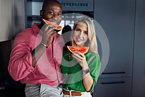 Multiracial couple eating melon in kitchen during hot sunny days.