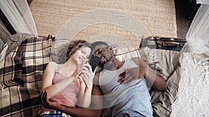 Multiracial couple lying on bed. Man and woman spend time together with joy and fan, use Smartphone, surf the internet.