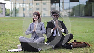 Multiracial coworkers, sitting on green grass outside, and doing yoga meditation in namaste pose