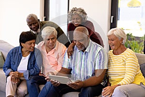 Multiracial curious seniors looking at male friend using digital tablet while sitting on sofa