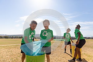 Multiracial elementary schoolboys putting garbage bags in garbage bin on school ground against sky