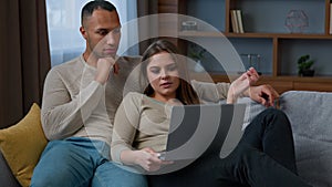 Multiracial ethnic couple homeowners shopping online use laptop hug on sofa at home diverse woman wife and man husband