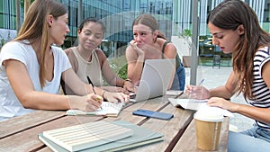 Multiracial female students studying and doing homework using laptops, laughing, talking outdoor in college campus.