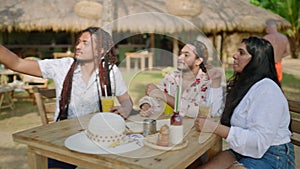 Multiracial friends enjoy drinks at tropical resto. Group laughs, chats, takes selfie. Afro man with braids captures