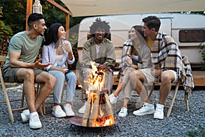 Multiracial friends sitting near bonfire, talking to each other, laughing, spending autumn evening near motorhome